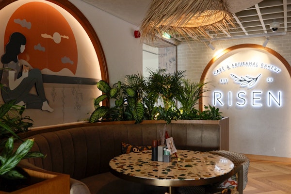 Experience the cozy ambiance at Risen Café & Artisanal Bakery. A stylish seating area, featuring greenery, a mural, and warm lighting.