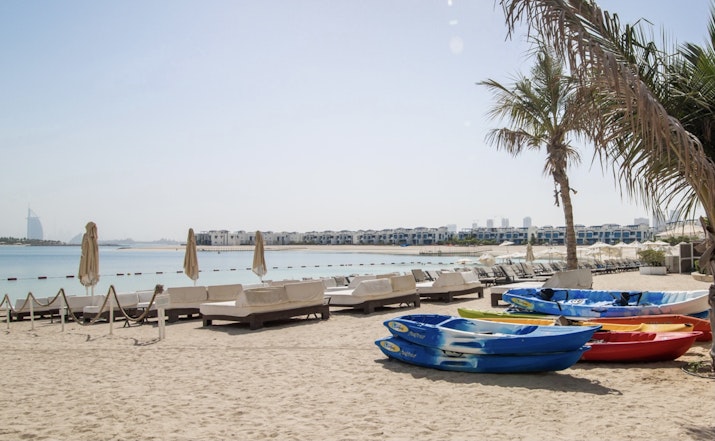 RIVA Beach Club A beach scene with colorful kayaks on the sand and lounge chairs, palm trees, and a calm waterfront.