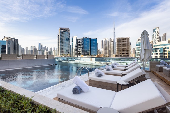 A rooftop pool with lounge chairs overlooking a city skyline, including the Burj Khalifa.
