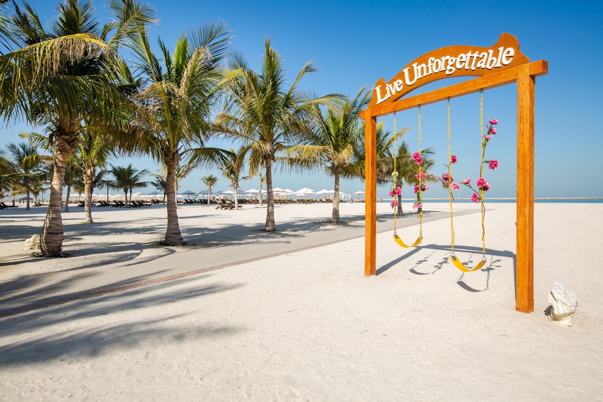 A swing set on the beach under the sign "Live Unforgettable," surrounded by palm trees and sand.