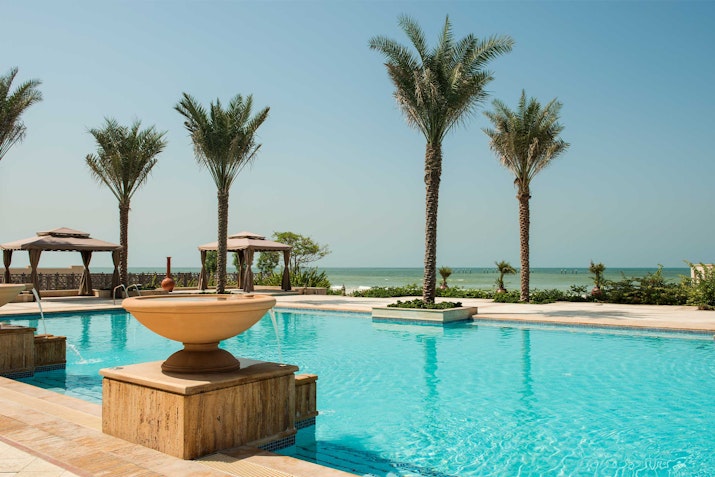 A serene poolside view with a fountain in the foreground, surrounded by palm trees, cabanas, and the beach in the distance.
