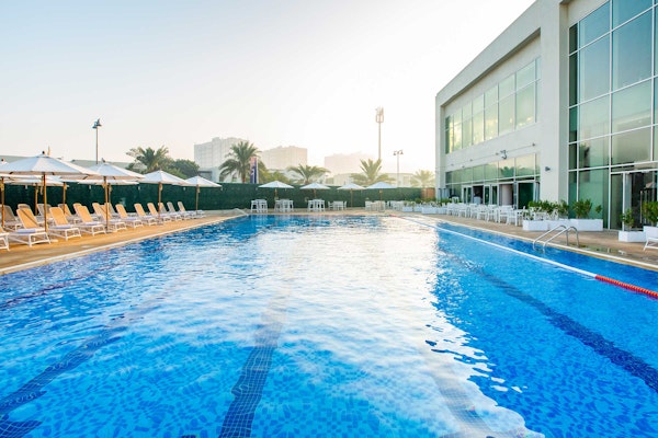 Swimming pool with lanes, surrounded by lounge chairs and a lifebuoy, with a building in the background.