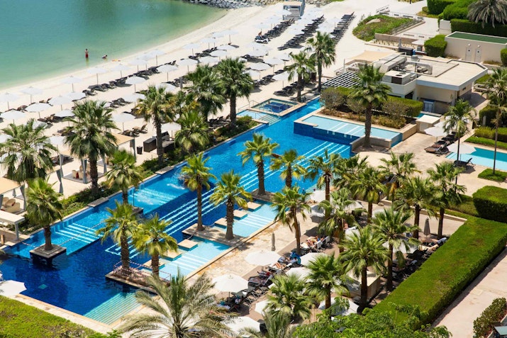 A luxurious pool area surrounded by palm trees, with sun loungers on one side and the beach in the distance. The pools are lined with geometric patterns and lush greenery.