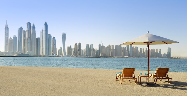 Beach with lounge chairs, umbrella, and Dubai skyline in the background.
