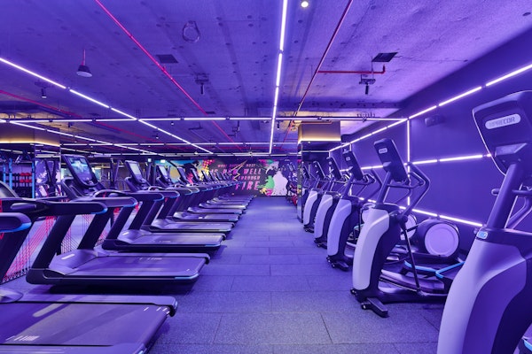 A modern gym interior featuring several treadmills lined up under vibrant purple and pink neon lights.