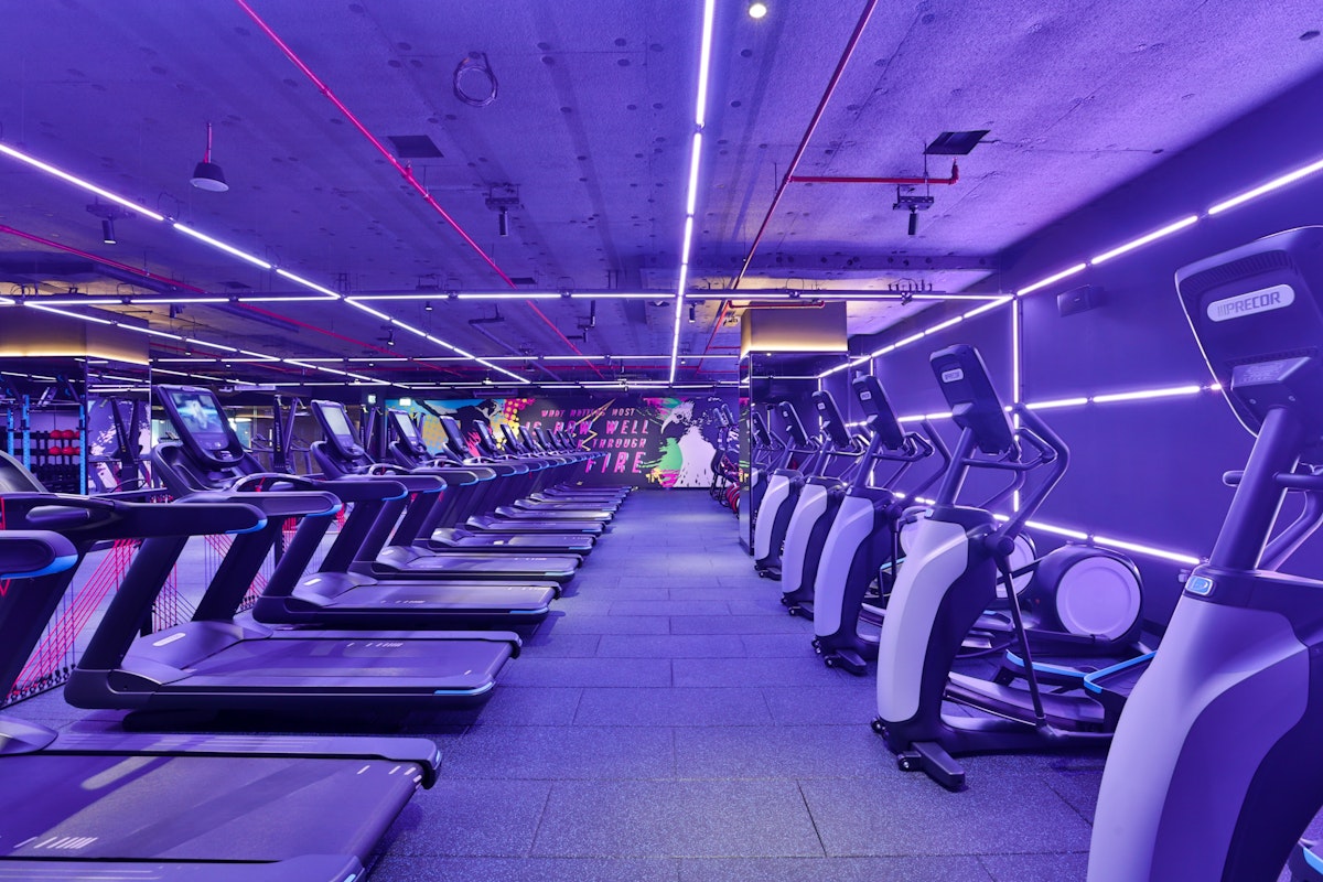 A modern gym interior featuring several treadmills lined up under vibrant purple and pink neon lights.