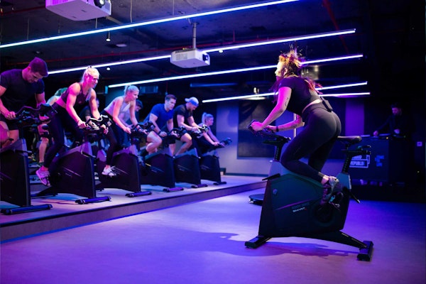 A group of people participating in a spinning class at a gym, with one woman in the foreground actively cycling and others in the background on stationary bikes, all under vibrant neon lights.