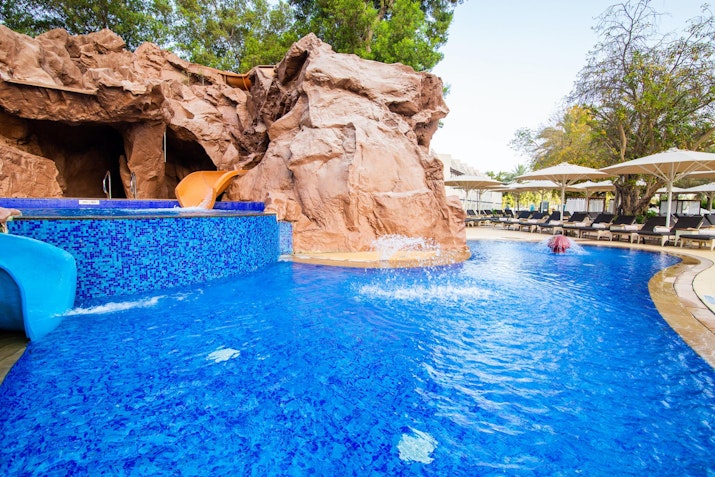 A swimming pool with a water slide, surrounded by a rocky structure and lounge chairs.