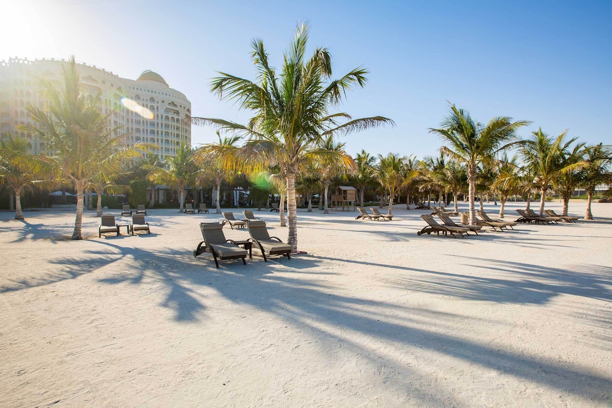A beach with lounge chairs under palm trees and clear blue skies, with the backdrop of a hotel building.