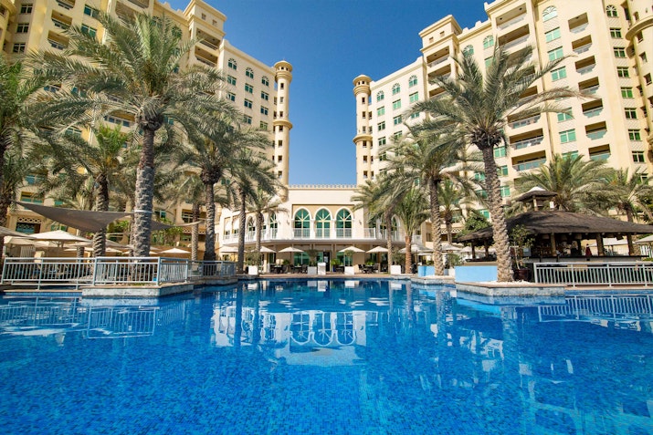 Luxury hotel pool surrounded by palm trees with a grand building in the background.