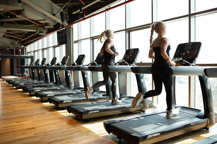 Two women running on treadmills in a bright gym with large windows.
