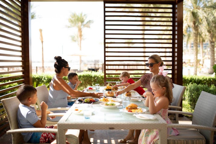 Sella at Dubai Polo Club A family enjoying a meal outdoors at a restaurant, with children and adults seated at a table, eating food under a shaded area surrounded by palm trees.