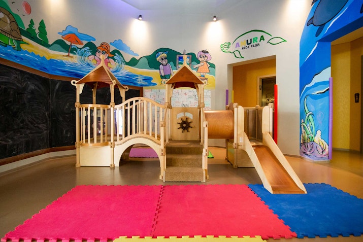 Children's play area with a wooden play structure, slide, and colorful floor mats, surrounded by vibrant murals.
