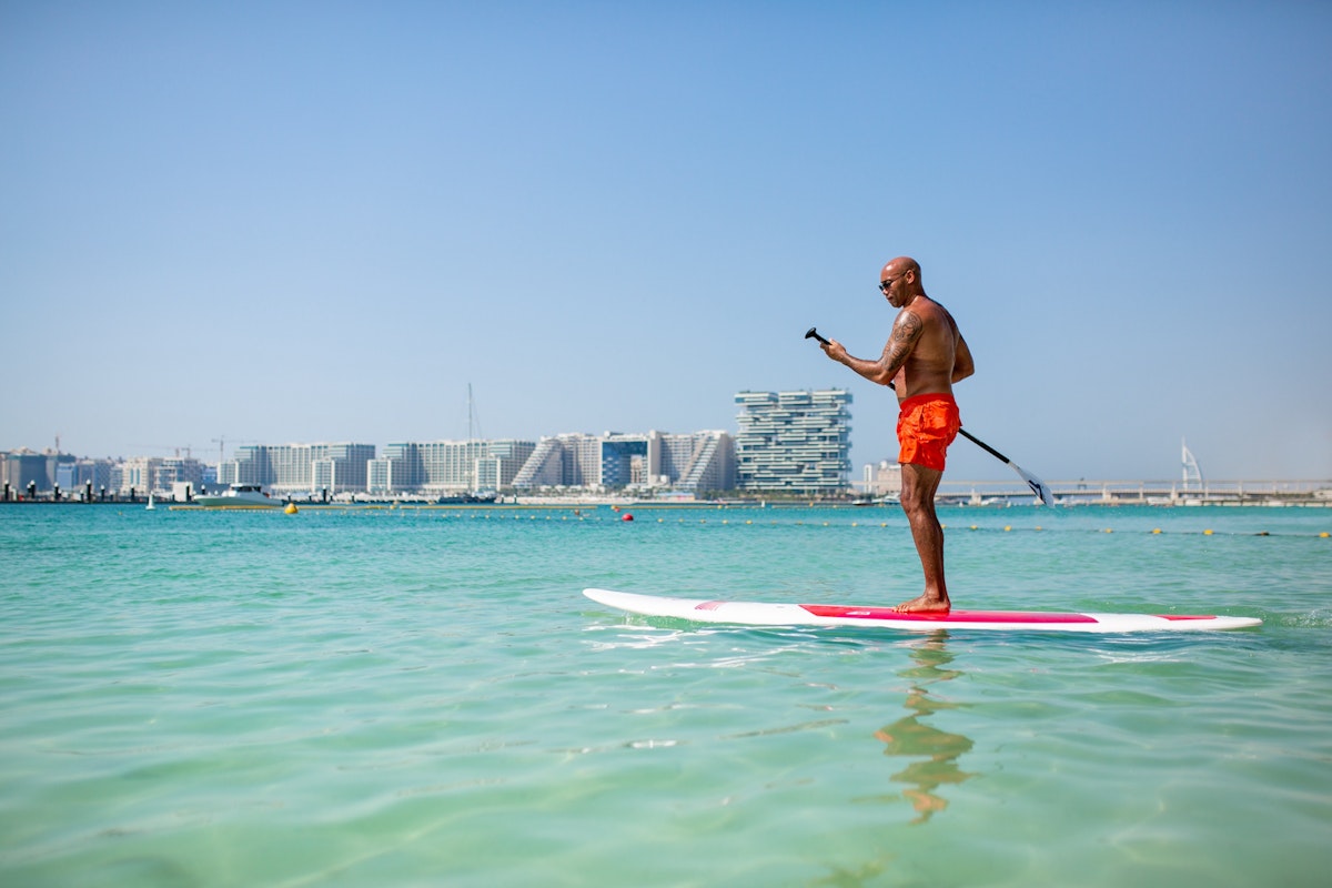 Man paddleboarding on clear turquoise water with cityscape in the background.