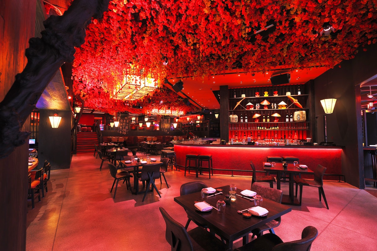 A luxurious restaurant with a red floral ceiling and ambient lighting, featuring dark wood tables and chairs, and a well-stocked bar in the background.