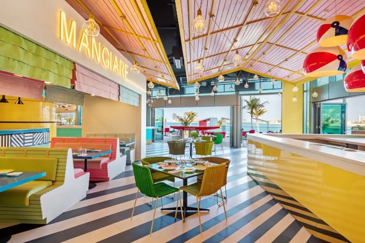 A vibrant and colorful restaurant interior with striped floors, playful light fixtures, and eclectic seating in green, yellow, and pink, with a neon "MANGIARE" sign on the wall.
