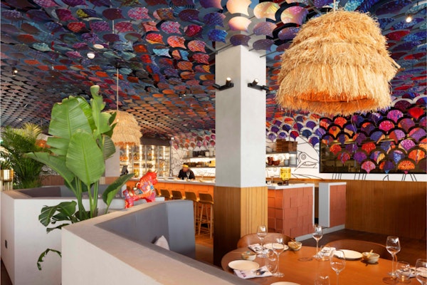 PLAYA Dubai A vibrant restaurant interior featuring colorful fan-shaped ceiling decor and woven hanging lights, with a cozy dining space and tropical plants.