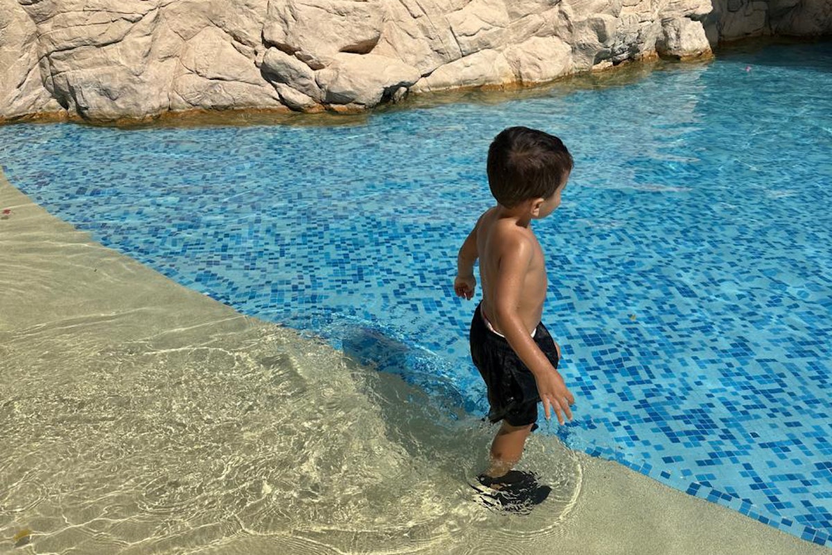 Top Summer Tips from a Mum Child standing at the edge of a pool with tiled blue water and rock backdrop.