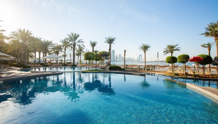 Fairmont The Palm A clear blue pool surrounded by palm trees and lounge chairs, with a view of a city skyline in the background.