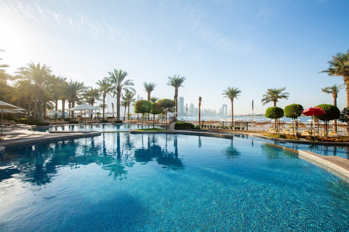 A clear blue pool surrounded by palm trees and lounge chairs, with a view of a city skyline in the background.
