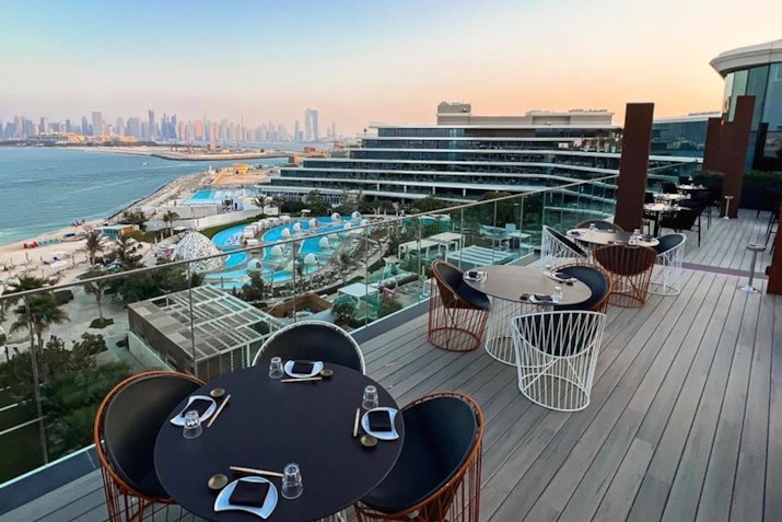 Rooftop dining area with tables overlooking a pool and a view of the city skyline.