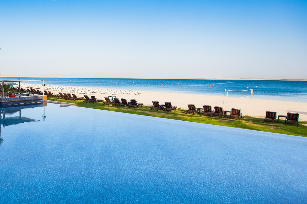 JA The Resort Serene infinity pool overlooking a sandy beach with lounge chairs and palm trees under a clear blue sky