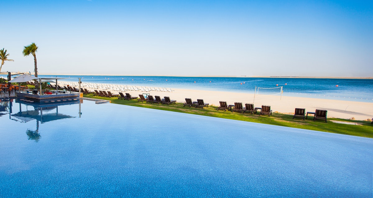 Serene infinity pool overlooking a sandy beach with lounge chairs and palm trees under a clear blue sky