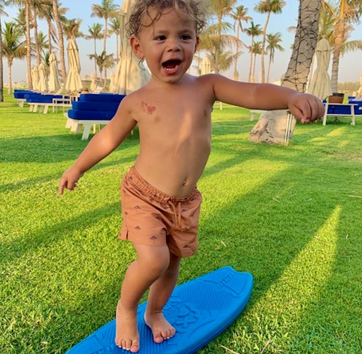 JA The Resort A joyful toddler standing on a blue surfboard on a grassy area, with palm trees and lounge chairs in the background