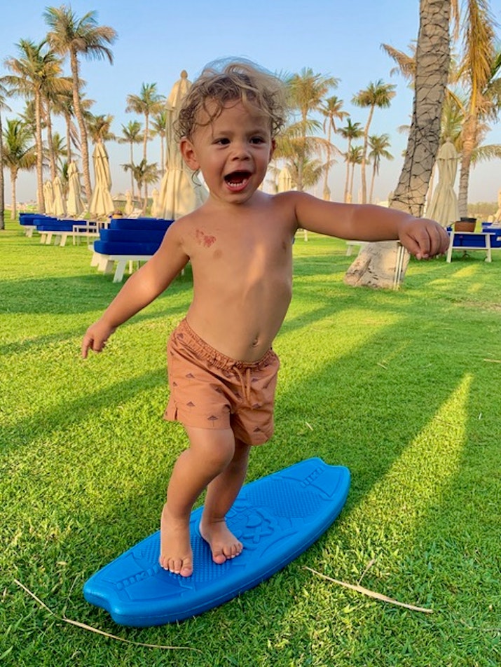 A joyful toddler standing on a blue surfboard on a grassy area, with palm trees and lounge chairs in the background