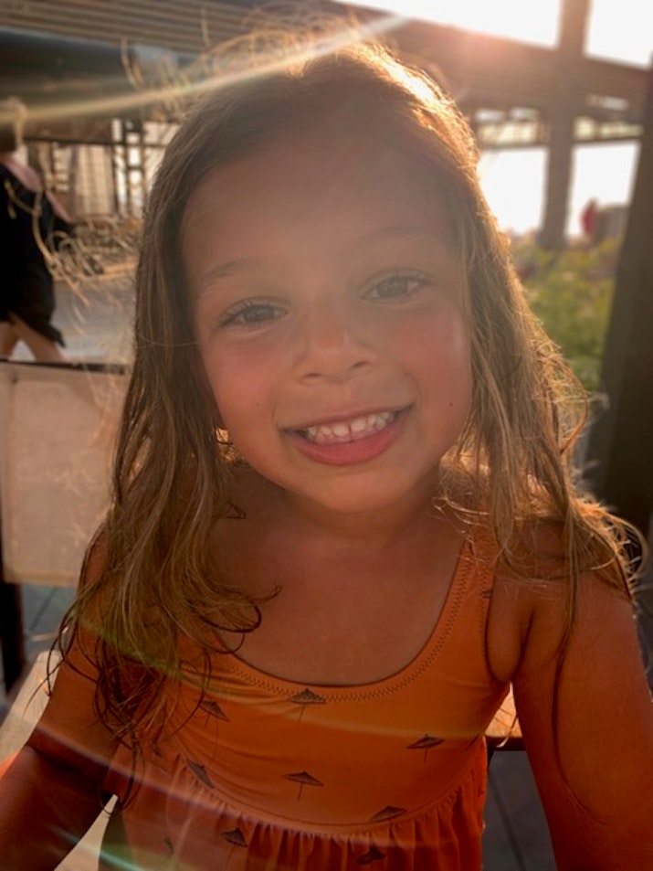 A smiling young girl with long hair, wearing an orange dress, captured in sunlight