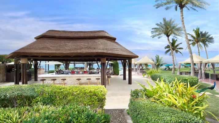 A thatched-roof beach hut surrounded by lush greenery and palm trees, with a view of the beach in the background.