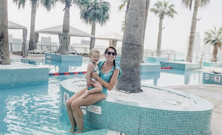 Mother and child sitting together in a pool, surrounded by palm trees.