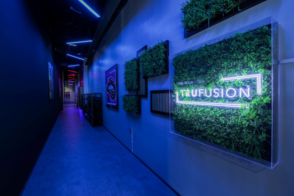 A modern hallway with neon blue lighting and greenery accents, featuring a prominent "Trufusion" sign on the wall.