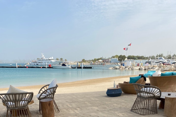 A tranquil beachside view featuring wicker seating, sandy shores, and boats docked in the calm water under a cloudy sky.