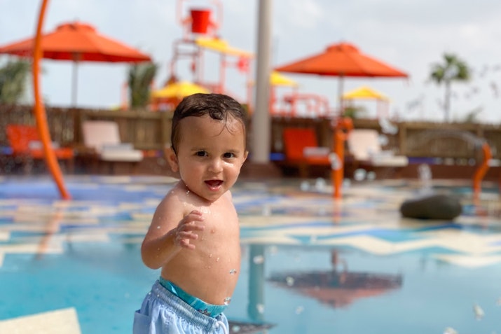 JA Beach Resort A cheerful toddler splashing in a water play area, wearing swim shorts and smiling at the camera.