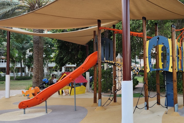 Colorful children's playground featuring a red slide and climbing structures under a shaded area.
