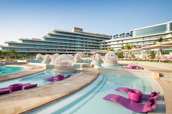 A luxurious pool area with modern purple lounge chairs and bubble-shaped pods, adjacent to a sleek hotel building.