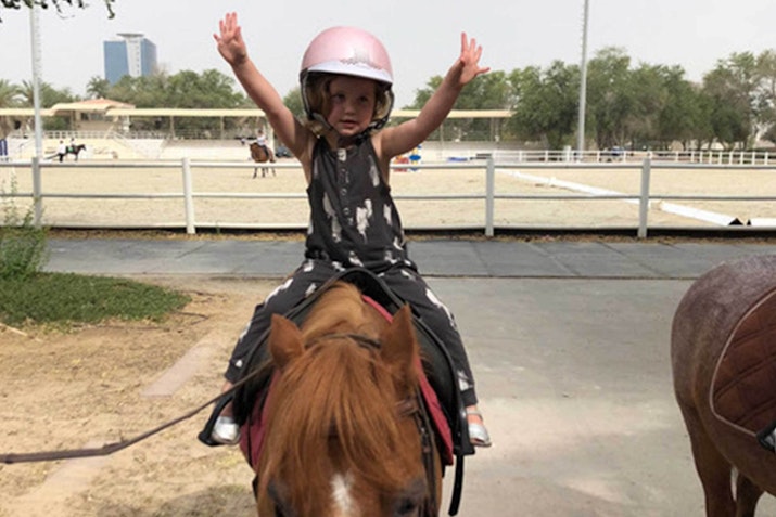 A child wearing a helmet and raising both hands while riding a pony at a horse riding facility.