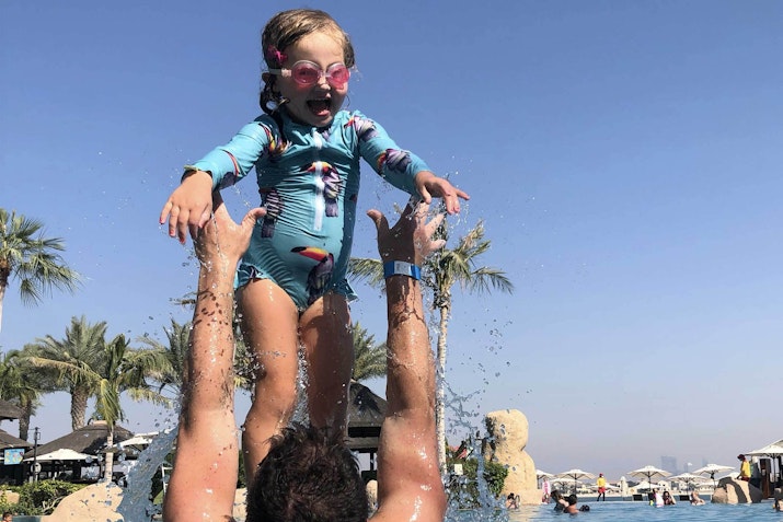 A father lifting his daughter above the water playfully while she enjoys the moment in a pool.