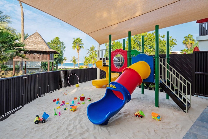 JA The Resort A colorful playground featuring a blue slide, yellow climbing structure, and various toys scattered on the sandy ground, all under a shaded area