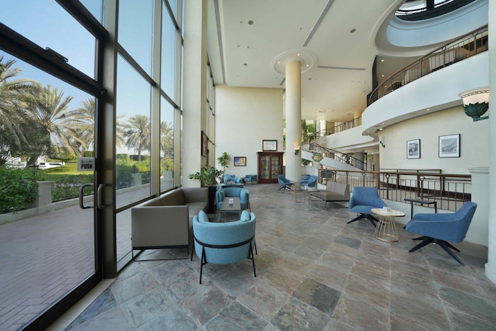 A bright and spacious hotel lobby with comfortable modern seating, large windows offering views of palm trees, and a stylish staircase leading to the upper floor.