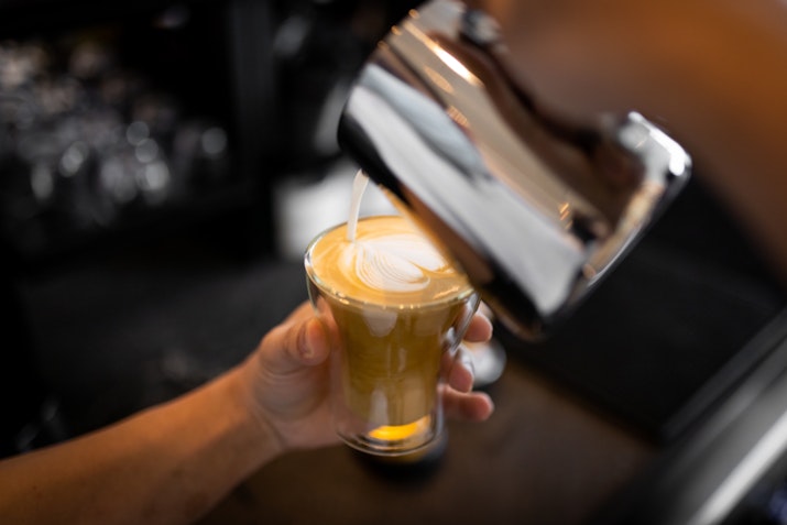 A person is pouring milk into a glass of coffee, creating a layered effect with the milk and espresso.