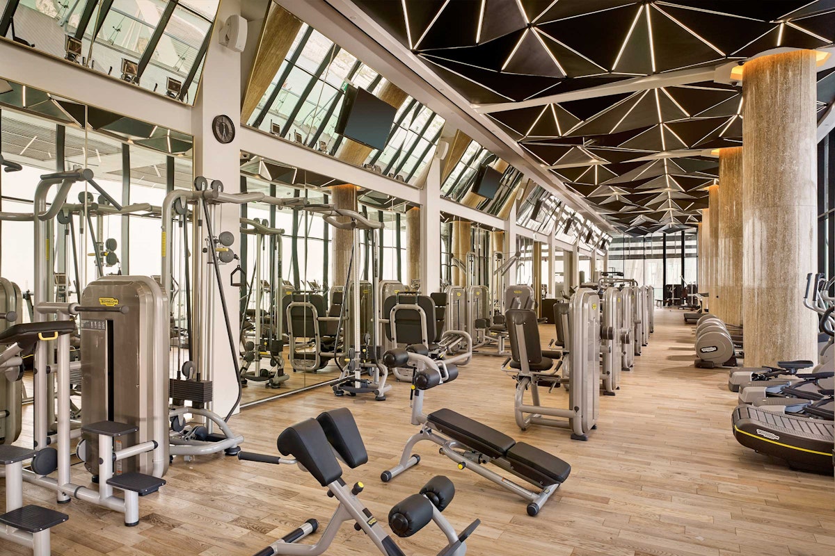 A modern gym with various exercise machines, natural light streaming through large windows, and a sleek geometric ceiling design.