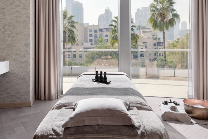 A serene spa treatment room featuring a massage table dressed in soft linens, with a large window offering a view of palm trees and the city skyline in the background, enhancing the calming atmosphere.