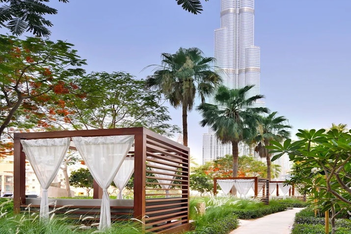 A tranquil outdoor area featuring wooden cabanas with white curtains, lush greenery, and tall palm trees, with the Burj Khalifa visible in the background against a clear sky.