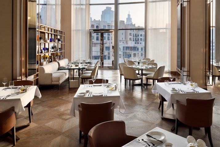 A sophisticated restaurant interior featuring elegantly set tables, modern furnishings, and large windows that allow natural light to fill the space, providing a view of the city skyline.