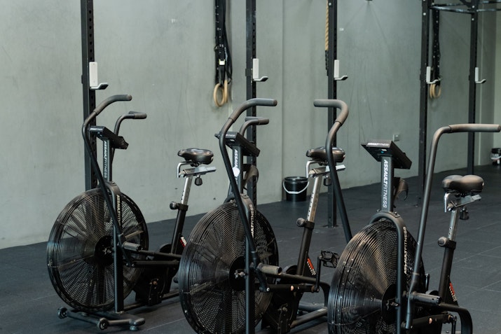 Close-up of a row of air bikes in a gym, positioned in front of a wall rig with gymnastic rings and resistance ropes, ideal for intense cardio workouts