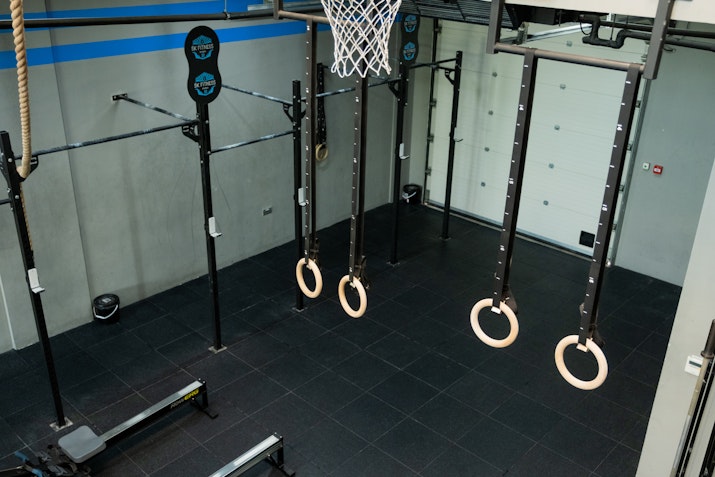 A gym area featuring gymnastic rings, ropes, pull-up bars, and a basketball hoop set against a clean, industrial backdrop with black tiled flooring.