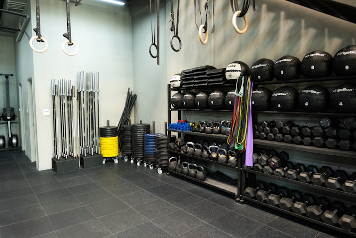 SK Fitness Gym storage area with neatly organized barbells, medicine balls, resistance bands, and dumbbells, set against a modern black-tiled floor with CrossFit rings hanging from the ceiling