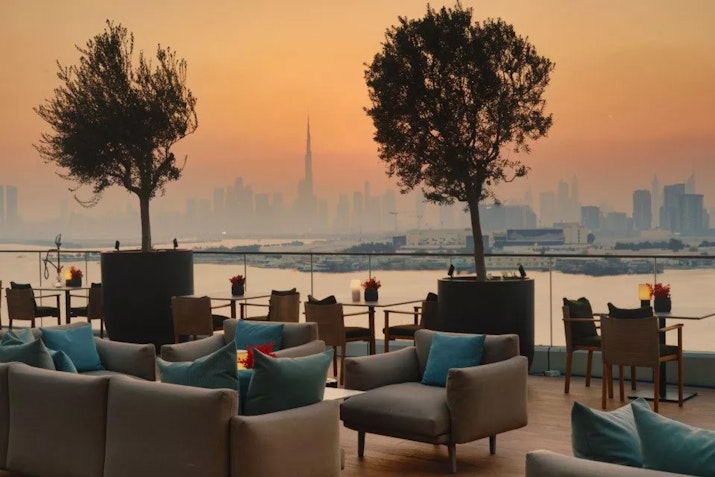 An elegant outdoor lounge area with comfortable seating and tables set against a sunset backdrop, overlooking a hazy city skyline, including the silhouette of a tall tower. The sky is painted in warm orange and pink hues.